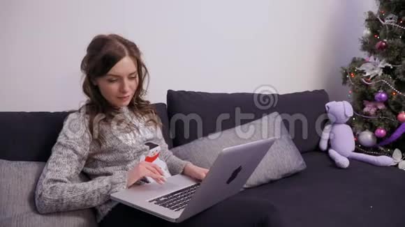 美丽的黑发女郎使用笔记本电脑坐在圣诞树附近的沙发上视频的预览图