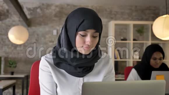年轻的穆斯林妇女戴着头巾在笔记本电脑上工作和打字两名妇女坐在办公室其中一名是非洲妇女视频的预览图
