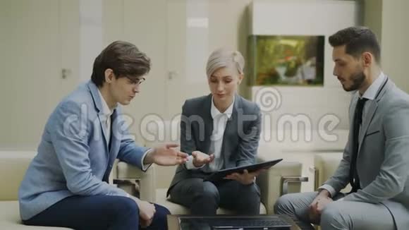 在现代的沙发上坐着的女商人用数码平板电脑与男商务同事展开了一场闹剧视频的预览图