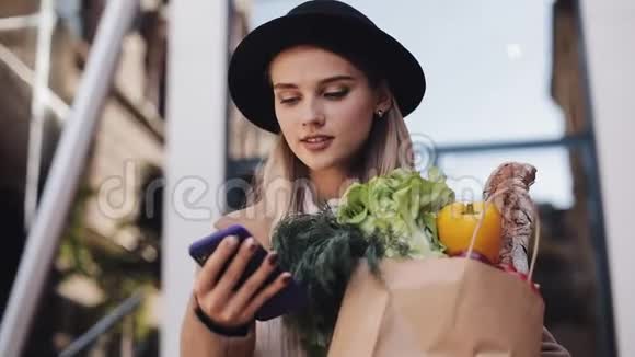 年轻漂亮的女人穿着时髦的外套站在街上拿着一包产品用智能手机说话视频的预览图