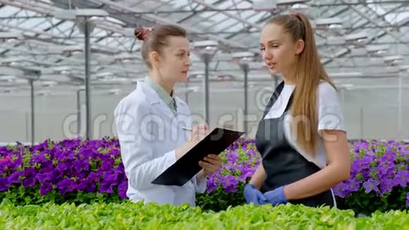 两个穿着白大褂黑围裙的女人科学家生物学家或农学家检查和分析花卉和绿色植物视频的预览图