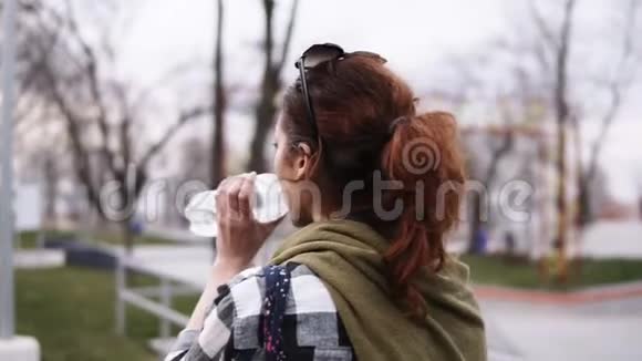 一个有尾巴的女孩在公园阴天散步从塑料瓶里喝水从背面看模糊的视频的预览图