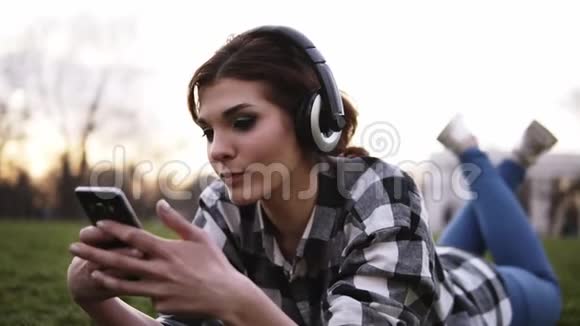 年轻女孩戴着时尚耳机躺在草地上听音乐在手机里聊天关门视频的预览图
