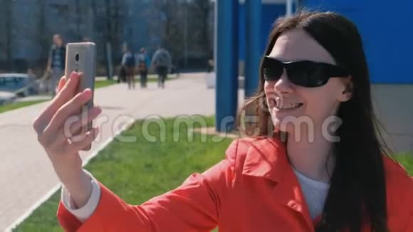 图为戴着墨镜的快乐的年轻黑发女子在蓝色大楼旁打电话视频的预览图