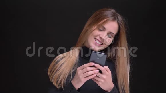 年轻的白种人长发金发金发女孩在黑色背景下微笑地看着智能手机的肖像视频的预览图