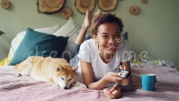 美国黑人妇女正在看电视手里拿着遥控器按着按钮一边笑着一边躺着她的宠物狗视频的预览图