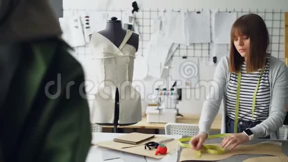 漂亮的女裁缝正在她的小裁缝店里用胶带测量衣服图案每天的工作视频的预览图