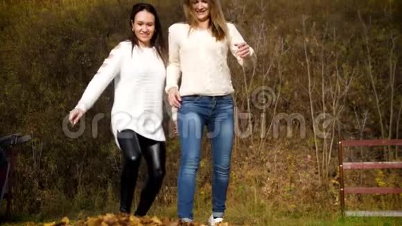 两个年轻女子将干枯的落叶踢向镜头两个女孩玩黄色的秋叶惹人的视频的预览图