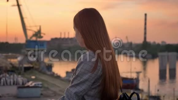 年轻的姜女穿着夹克站在桥上看着河边的工厂夕阳西下视频的预览图