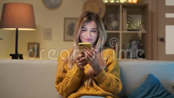 金发女郎坐在沙发上用手机聊天在一个舒适的房间里大笑的肖像室内视频的预览图