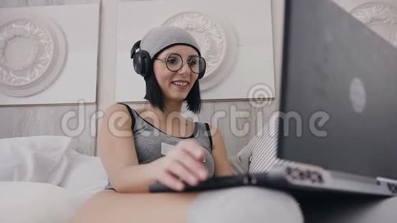 戴着黑色耳机和灰色帽子的漂亮女孩或女人听着音乐在笔记本电脑上工作视频的预览图