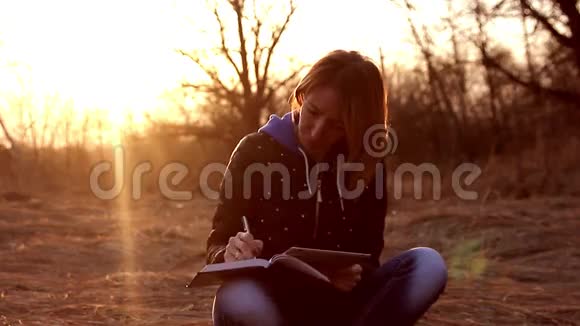 美丽的少女坐在公园秋天落叶上读着平板电脑在笔记本上做笔记视频的预览图