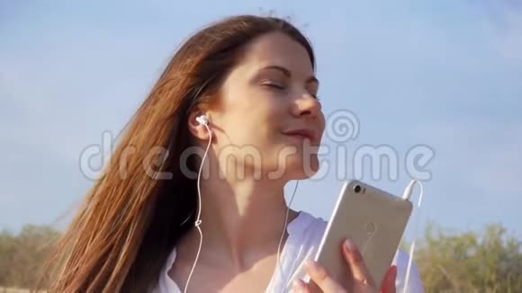 年轻快乐的女人用手机对抗蓝天女性通过手机慢动作听音乐视频的预览图