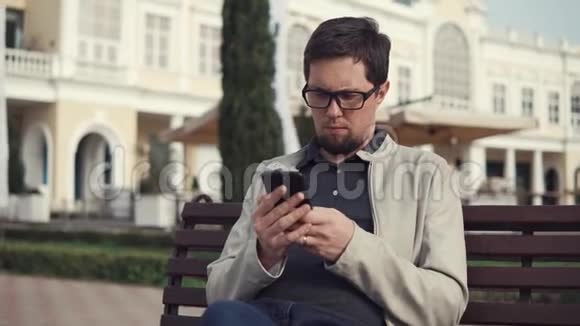 一个戴眼镜的严肃男人在公园的长椅上用智能手机打印短信视频的预览图