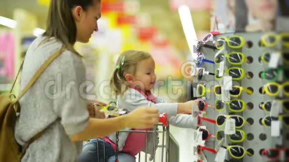 年轻的妻子穿着休闲服装为她的小女儿选择太阳镜站在超市货架附近视频的预览图