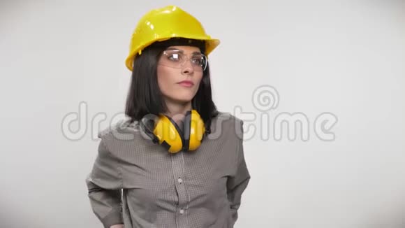年轻建筑工人妇女戴着耳机眼镜和头盔双手交叉站立表情严肃白色背景视频的预览图