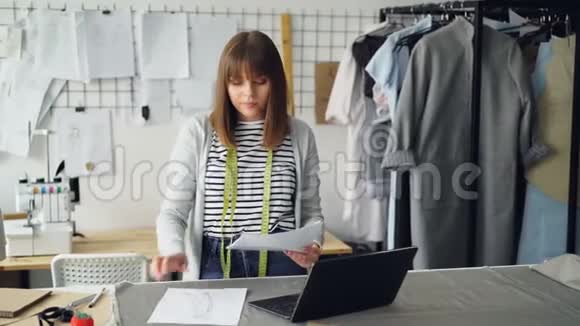 年轻有魅力的女人正在为新的时装秀选择草图并在她的裁缝店笔记本电脑创意创意视频的预览图