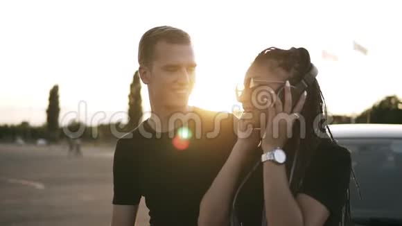 漂亮的年轻女士戴着时尚的太阳镜和男朋友一起在街上用耳机听音乐视频的预览图