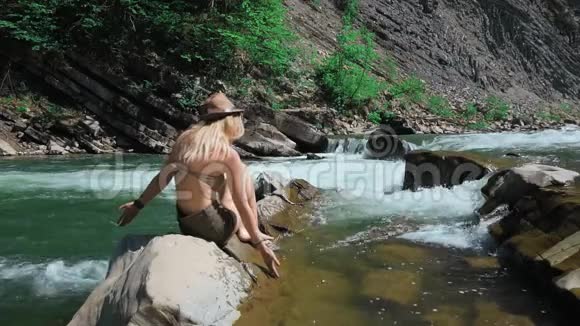 一位戴着帽子的年轻女子坐在瑞士的一条山河的岸边他用手摸着水视频的预览图
