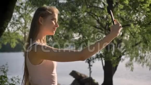 一位留着长发的年轻美女带着手机微笑着自拍停在河边背景很美视频的预览图