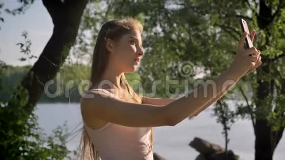 年轻漂亮的女人拿着手机自拍对着镜头微笑站在河边的公园里视频的预览图
