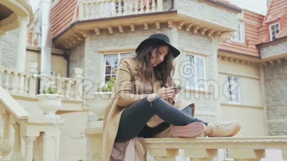 迷人的欧洲微笑卷发戴着时尚的帽子和外套在她的智能手机上和某人交谈视频的预览图
