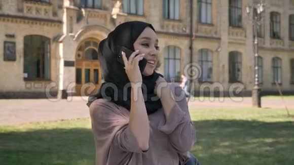 戴着头巾的年轻可爱的穆斯林女孩正在电话中交谈在夏天的白天笑着并在背景中建立起来视频的预览图