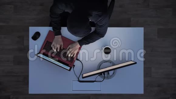 穿着黑色连帽衫的年轻人坐在办公桌后在笔记本电脑上打字然后在夜间亮灯时逃跑视频的预览图