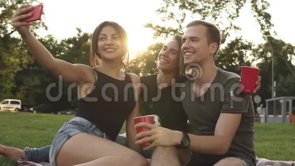 三个朋友坐在公园的草地上摆姿势拍照的肖像布鲁内特女孩和她一起拍照视频的预览图