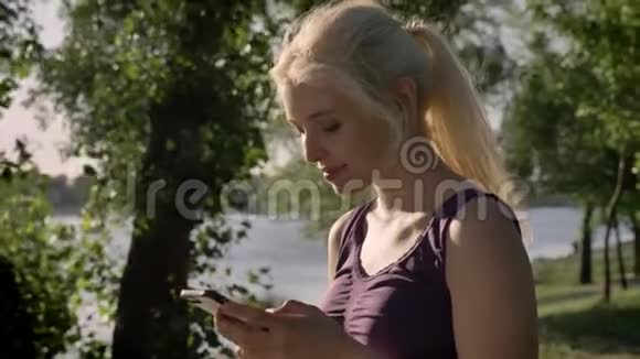 年轻的美女金发在手机上发短信看着镜头微笑着停在河边的背景下视频的预览图