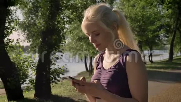 年轻的金发女郎梳着马尾辫在手机上打字看着镜头微笑着停在河边的背景附近视频的预览图