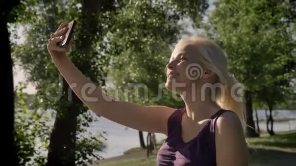 年轻漂亮的金发女郎用手机自拍在河边的公园里露出舌头和微笑视频的预览图