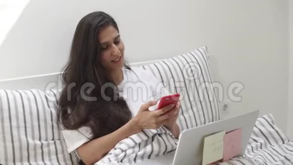 年轻女子在手机上打电话给朋友女士躺在笔记本电脑上视频的预览图
