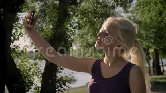 金发美女带着手机带着手机微笑着自拍停在河边的背景下视频的预览图