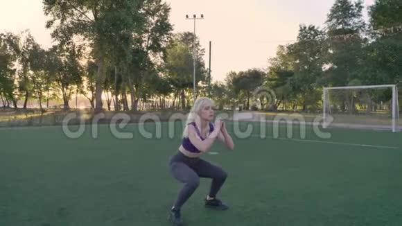 年轻的金发女人在公园的足球场上做蹲跳锻炼早上锻炼视频的预览图