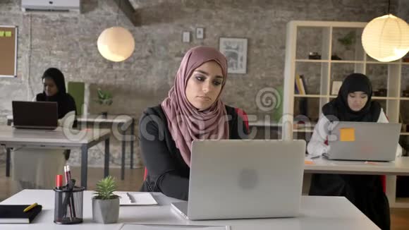 三个年轻的穆斯林妇女戴着头巾坐在现代办公室的笔记本电脑上打字穆斯林妇女打哈欠和工作视频的预览图