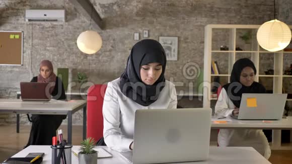 三位年轻的穆斯林妇女戴着头巾坐在现代办公室工作漂亮的女工在笔记本电脑上打字视频的预览图