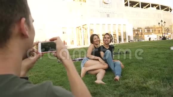 年轻的高加索男人带着智能手机为他的两个美丽的女性朋友拍照女孩们坐在绿色的地上视频的预览图