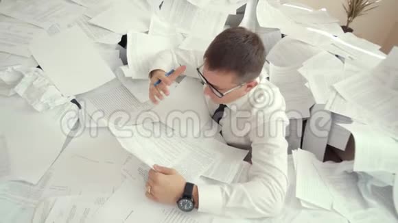 一个戴眼镜的年轻疲惫的人坐在一堆文件里抱着他的头由于文件数量一个办公室视频的预览图
