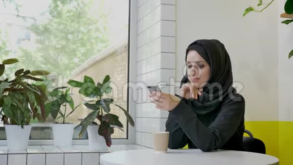 年轻漂亮的穆斯林女人戴着头巾拿着电话在手机上聊天微笑着坐在咖啡馆里快乐着视频的预览图