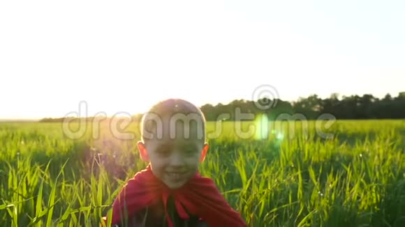 一个穿着超级英雄服装穿着红色斗篷的快乐孩子坐在一片绿色的草坪上映衬着夕阳然后升起视频的预览图