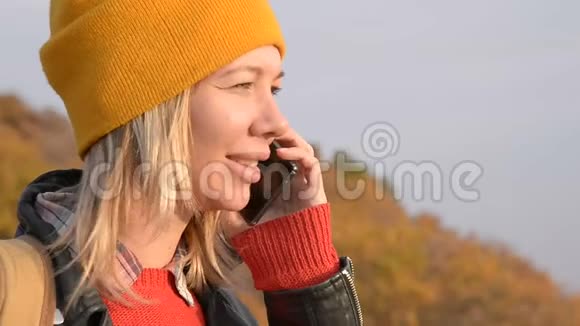 秋天一个戴着黄色帽子的漂亮白种人女孩正在户外用手机聊天微笑的女孩视频的预览图