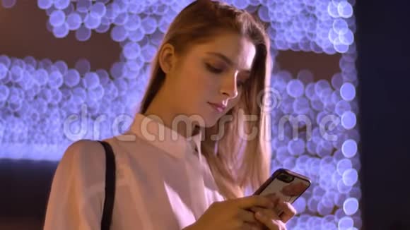 年轻可爱的女孩在夏天晚上在她的智能手机上打字交流的概念背景灯视频的预览图