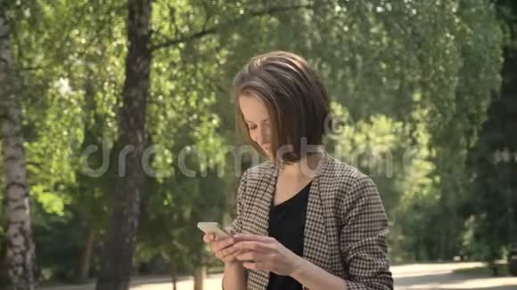 年轻迷人的集中注意力的女孩白天在公园里的智能手机上输入信息夏天在公园里看着摄像机视频的预览图