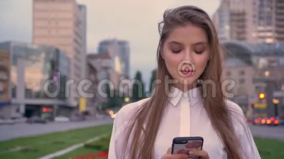 年轻漂亮的女孩正在城市中心的日落时分在智能手机上输入信息抚摸着头发微笑着视频的预览图