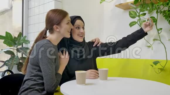 两个年轻美丽的穆斯林妇女坐在咖啡馆里其中一个戴着头巾自拍看着电话微笑着视频的预览图