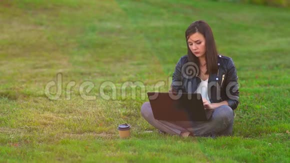 公园里的一个女孩在草坪上用笔记本电脑工作电话铃响接来电话视频的预览图