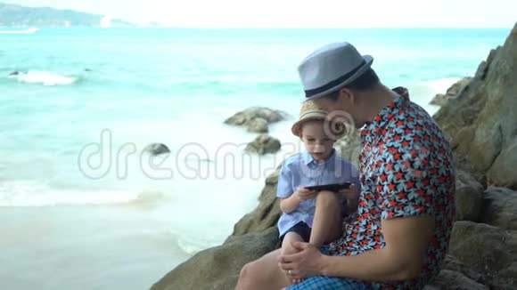 儿子和爸爸拿着电话坐在海边的岩石海岸上视频的预览图