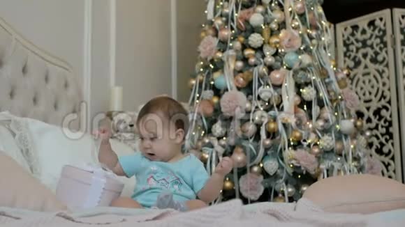一个小女孩正拿着礼物坐在圣诞树背景的床上视频的预览图