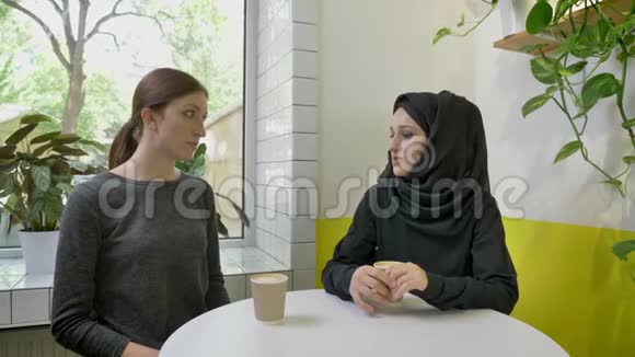 两个年轻漂亮的女人坐在咖啡馆里其中一个是戴着头巾的穆斯林女人跟其他女人说些什么视频的预览图